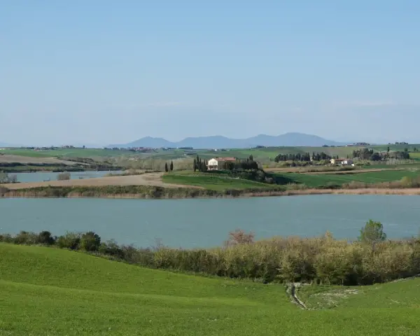 Campagna toscana con lago e casale in collina, un luogo dove la **santa luce cosa vedere** è ovunque, tra dolci pendii verdi e il cielo azzurro.