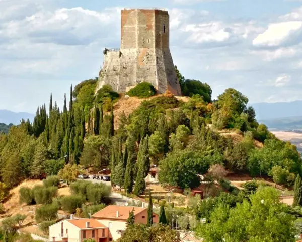 La rocca di Tentennano domina la collina, circondata da cipressi e ulivi. Sotto, un borgo con tetti rossi si adagia nel paesaggio toscano.