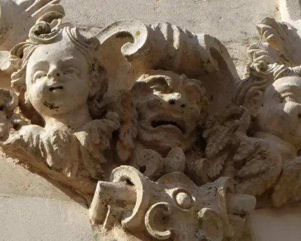 Dettaglio scultoreo barocco a Martina Franca: volti di putti e una maschera grottesca emergono dalla pietra calcarea, testimoni di un'antica arte.