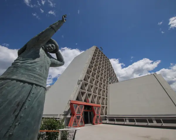 Statua bronzea davanti al Santuario Monte Grisa, con il suo ingresso rosso e la facciata a triangoli.