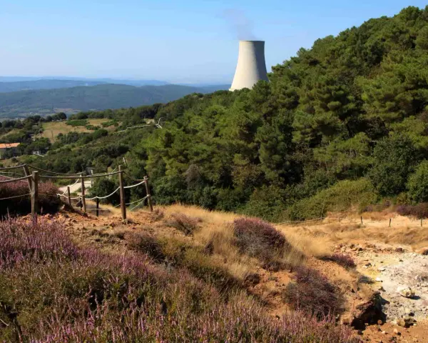 Sentiero nel parco delle Biancane con torretta di raffreddamento in lontananza, circondato da vegetazione e colline.