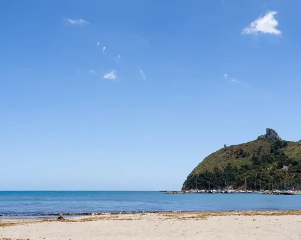 Spiaggia del Poetto con mare azzurro, montagne verdi e un porto turistico pieno di barche.