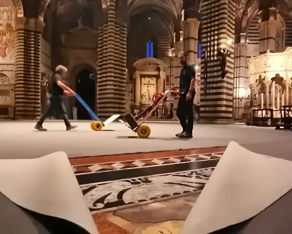 Due operai spostano carrelli sul magnifico pavimento del Duomo di Siena, mentre un telo protettivo è parzialmente sollevato, rivelando i dettagli artistici.