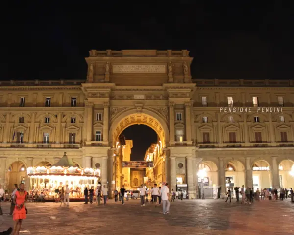 La piazza della Repubblica a Firenze di notte, con il suo arco monumentale illuminato, gente che passeggia e una giostra scintillante.