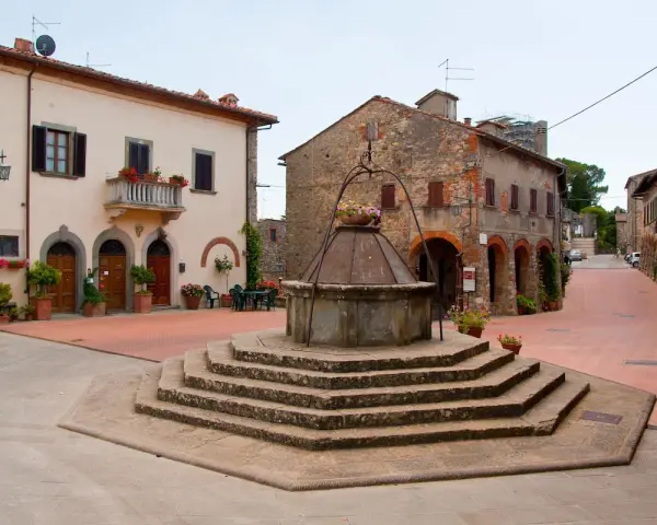 Piazza con pozzo antico a Civitella in Val di Chiana, circondata da case in pietra e intonaco, con fiori colorati.