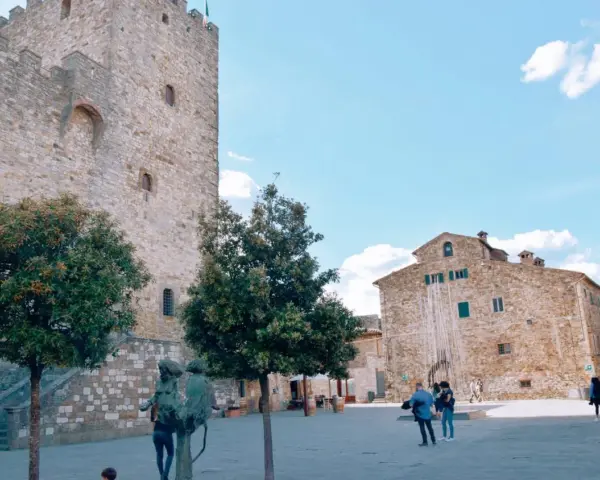 Piazza nel centro storico di Castellina in Chianti con torre medievale, edifici in pietra e scultura.