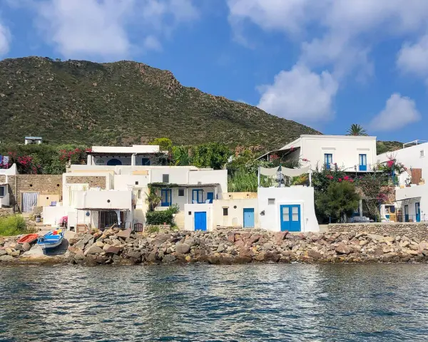 Case bianche con porte blu sull'isola di Panarea, con il mare calmo e una collina verde sullo sfondo.