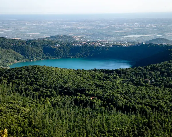 Vista panoramica del lago di Albano, circondato da rigogliosa vegetazione, con il borgo di Rocca di Papa in lontananza.