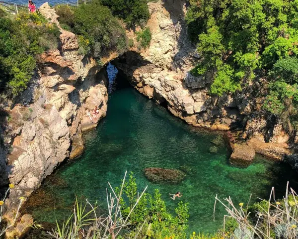 Bagni Regina Giovanna: una baia nascosta con acque cristalline, rocce scolpite e vegetazione lussureggiante. Persone si godono il sole e nuotano.
