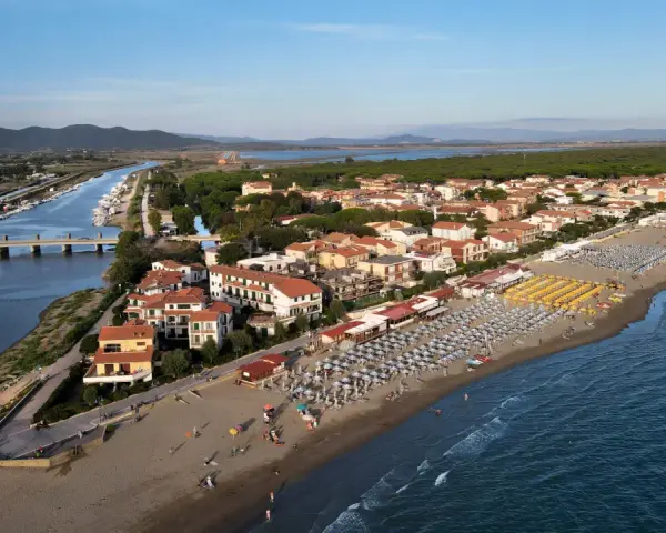 Vista aerea di Castiglione della Pescaia: un canale navigabile con barche, case con tetti rossi e una spiaggia attrezzata con ombrelloni e lettini, bagnata dal mare.