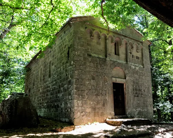 Antica pieve in pietra nel bosco Isabella, Radicofani. La luce filtra tra gli alberi illuminando la facciata.
