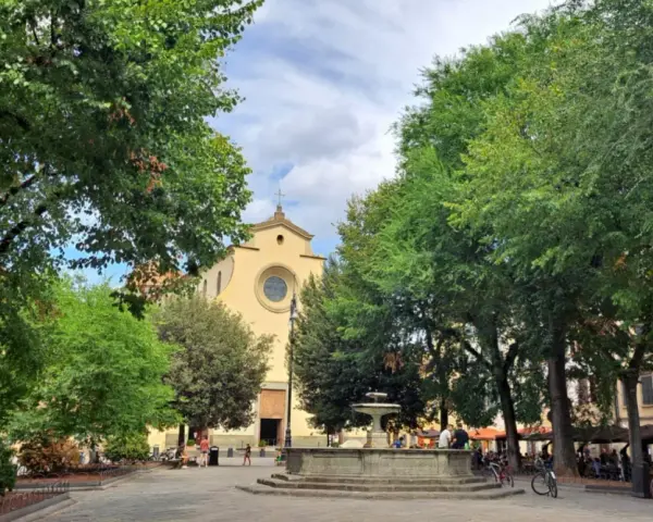 Piazza Santo Spirito a Firenze, con la chiesa sullo sfondo, una fontana al centro e alberi rigogliosi che incorniciano la scena.