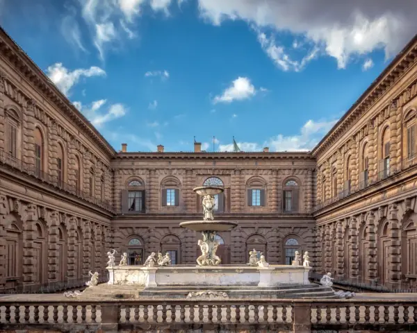 Cortile del Palazzo Pitti con fontana e statue, sotto un cielo azzurro con nuvole bianche.