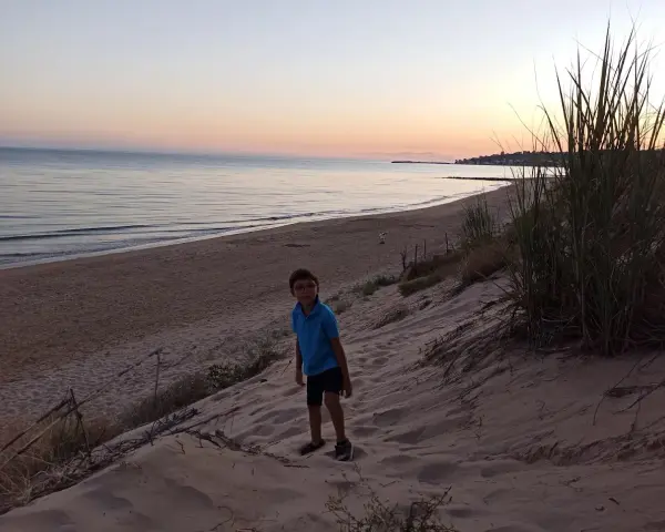 Bambino con occhiali su una duna di sabbia a Porto Palo, con il mare calmo e il cielo al tramonto sullo sfondo.
