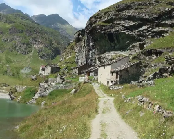 Antico borgo in pietra nel **Val di Susa**, con un sentiero che sale verso le case arroccate su un pendio verdeggiante, vicino a un lago alpino.