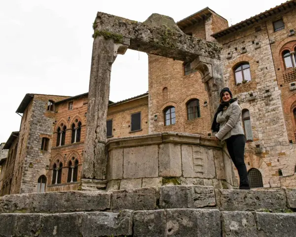 Donna sorride vicino all'arco in pietra e al pozzo di San Gimignano, con palazzi medievali sullo sfondo.