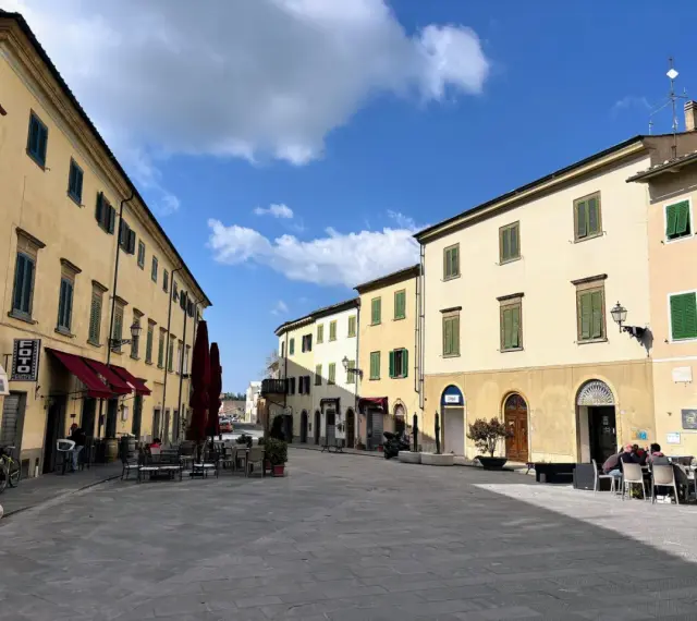 Piazza tranquilla a Pomarance, cosa vedere: edifici storici con persiane verdi, tavolini all'aperto di un bar.