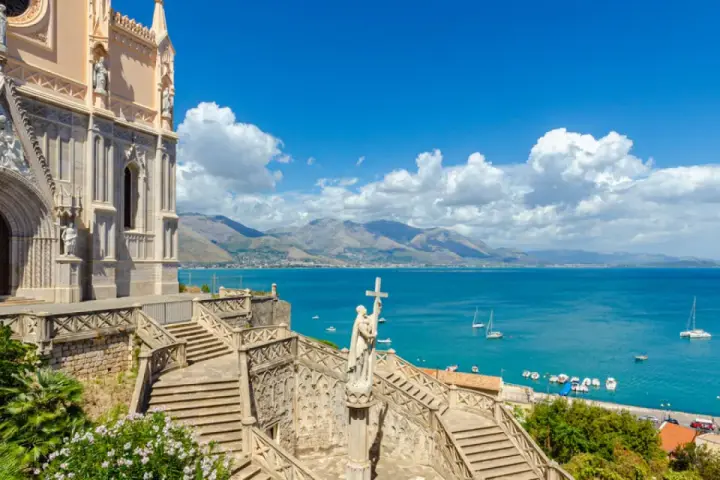 La chiesa domina il mare azzurro della Riviera di Ulisse. Scale imponenti conducono a un portale gotico, mentre barche solcano le acque.