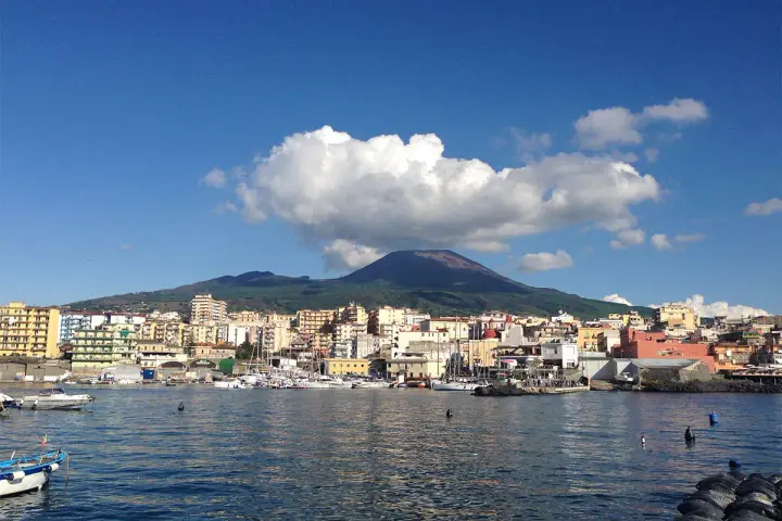 Vista del porto di Torre del Greco con il Vesuvio sullo sfondo, sotto un cielo azzurro con nuvole bianche.