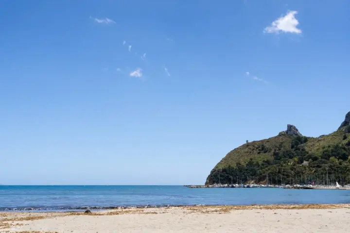 Spiaggia del Poetto con mare azzurro, montagne verdi e un porto turistico pieno di barche.