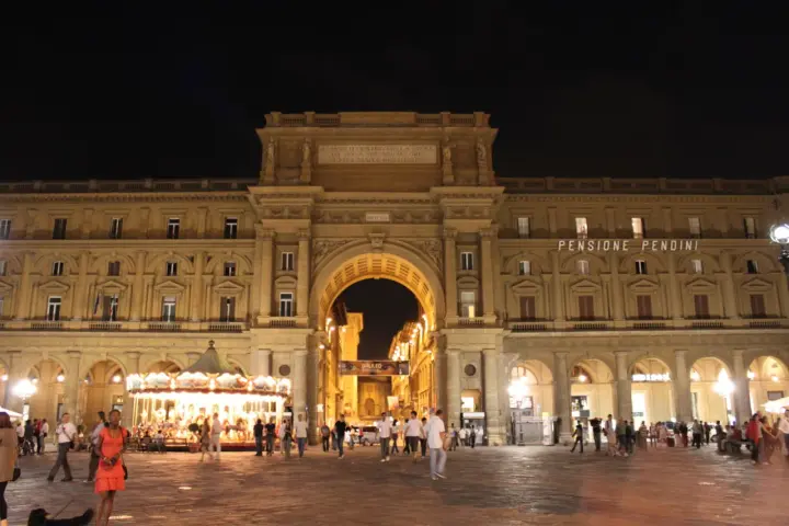 La piazza della Repubblica a Firenze di notte, con il suo arco monumentale illuminato, gente che passeggia e una giostra scintillante.