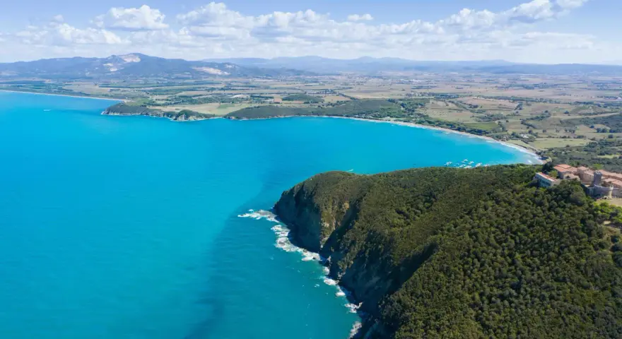 Vista aerea della costa degli Etruschi con mare azzurro, scogliere verdi e un borgo storico.