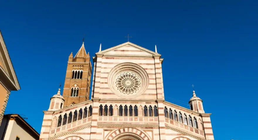 Il Duomo di Grosseto, con la sua facciata a strisce bianche e rosa e il rosone centrale, si staglia contro un cielo azzurro intenso.