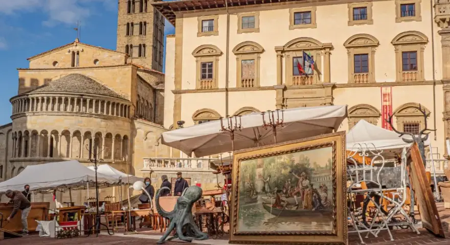 Mercatino d'antiquariato in piazza Grande ad Arezzo, con opere d'arte, sculture e mobili esposti sotto il sole.