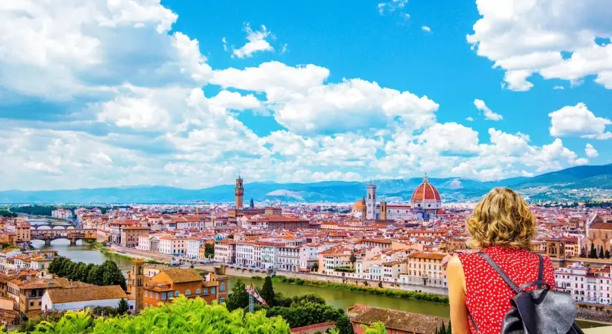 Vista mozzafiato di Firenze dal Piazzale Michelangelo. Una donna ammira il Duomo e il Ponte Vecchio sotto un cielo azzurro con nuvole bianche.