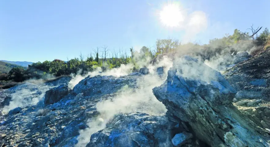 Fumarole attive nella valle del diavolo, con rocce grigie e blu avvolte dal vapore. Il sole splende nel cielo azzurro.