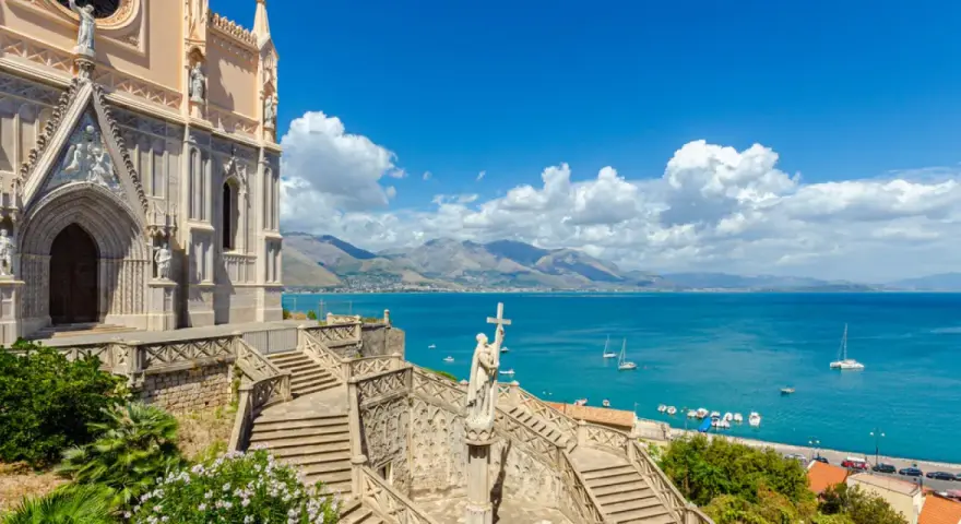 La chiesa domina il mare azzurro della Riviera di Ulisse. Scale imponenti conducono a un portale gotico, mentre barche solcano le acque.