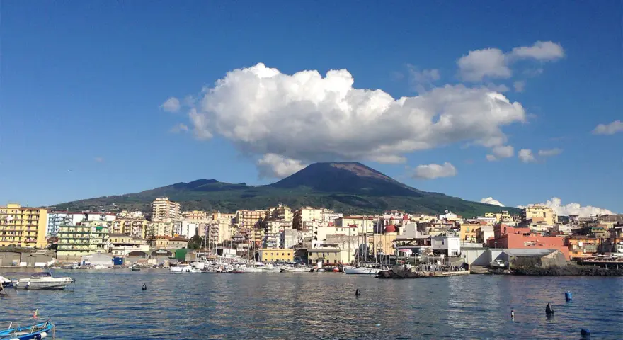 Vista del porto di Torre del Greco con il Vesuvio sullo sfondo, sotto un cielo azzurro con nuvole bianche.