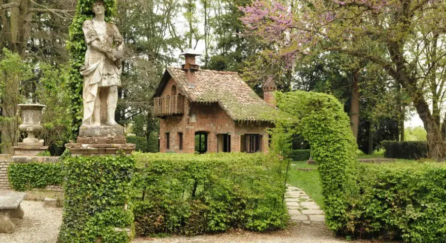 Giardino incantato a Grazzano Visconti con statua romana, casetta in mattoni e sentiero tra siepi fiorite.