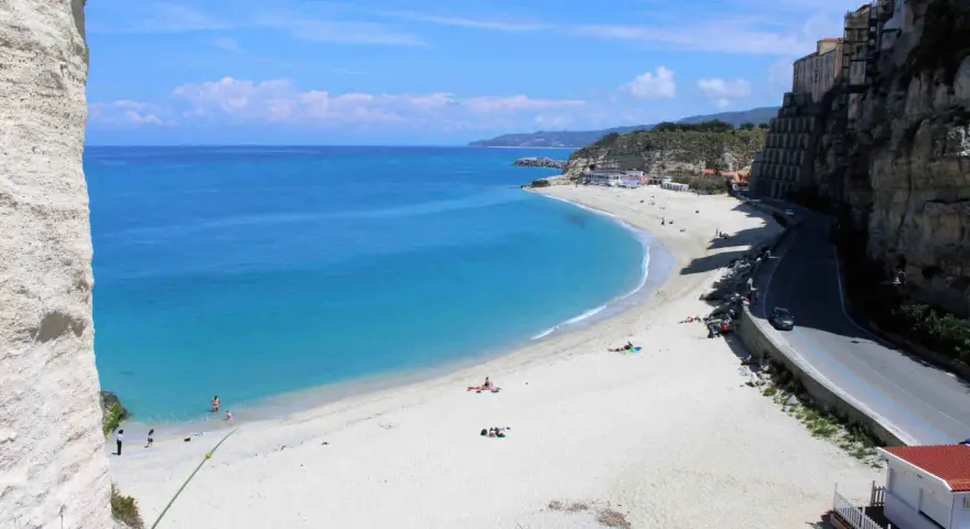 Spiaggia bianca e mare cristallino sulla **Costa degli Dei**. Case aggrappate alla scogliera, una strada panoramica e bagnanti che si godono il sole.