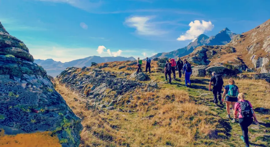 Un gruppo di escursionisti cammina su un sentiero roccioso e erboso, con montagne sullo sfondo. Il **Passo dello Spluga** offre panorami mozzafiato.