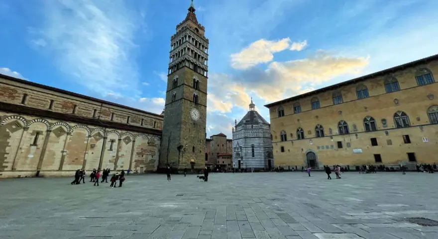 Piazza del Duomo di Pistoia con la torre campanaria, il battistero e il palazzo vescovile sotto un cielo nuvoloso.