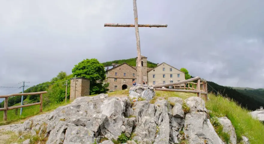 Croce di legno a San Pellegrino in Alpe, con edifici storici sullo sfondo e rocce in primo piano.