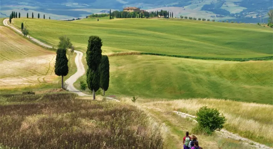 Sentiero tortuoso nel Val d'Orcia, con colline verdi, cipressi e un casale in lontananza. Una mappa ideale per un'escursione.