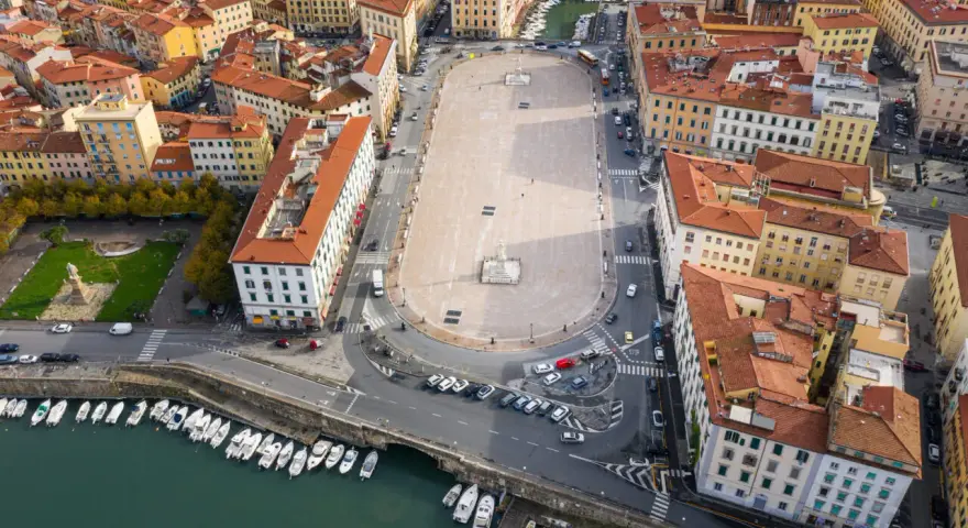 Vista aerea della **Piazza della Repubblica a Livorno**, con edifici storici, un monumento centrale, auto e barche ormeggiate nel canale.