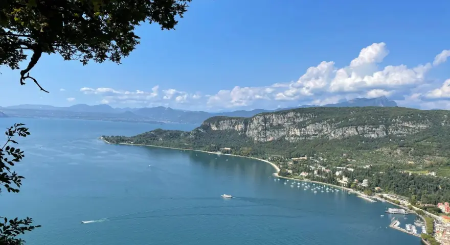 Vista panoramica del Lago di Garda con San Zeno di Montagna sullo sfondo, tra montagne verdi e cielo azzurro.