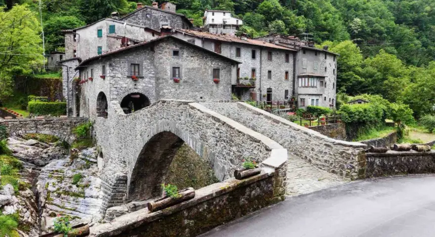 Ponte in pietra a Vergemoli, con case in pietra e vegetazione lussureggiante sullo sfondo. Le antiche fabbriche di Vergemoli sembrano emergere dalla collina.