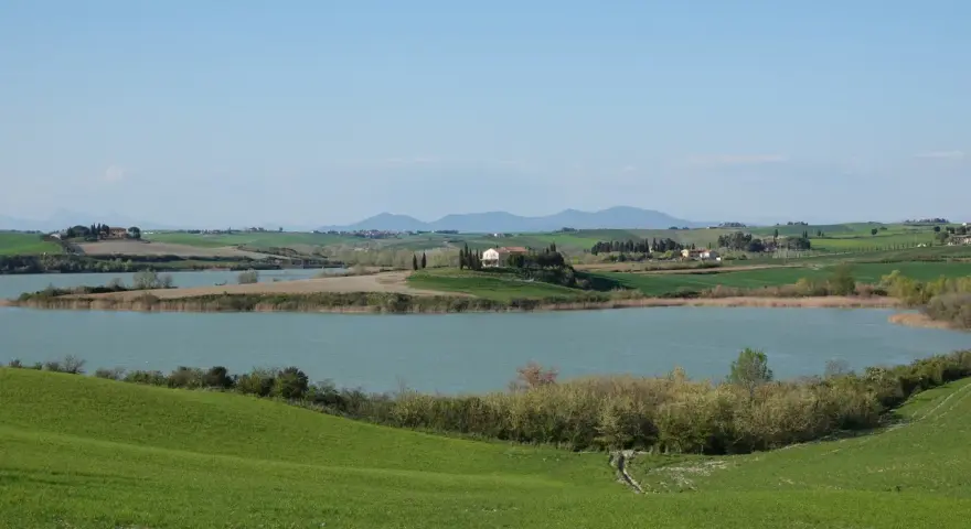 Campagna toscana con lago e casale in collina, un luogo dove la **santa luce cosa vedere** è ovunque, tra dolci pendii verdi e il cielo azzurro.