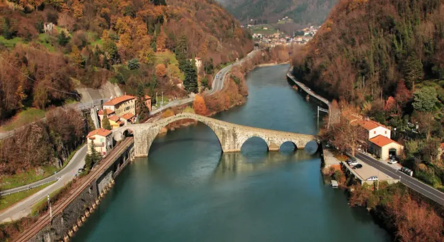 Il **ponte della Maddalena**, con i suoi archi irregolari, attraversa un fiume circondato da colline autunnali e case colorate.