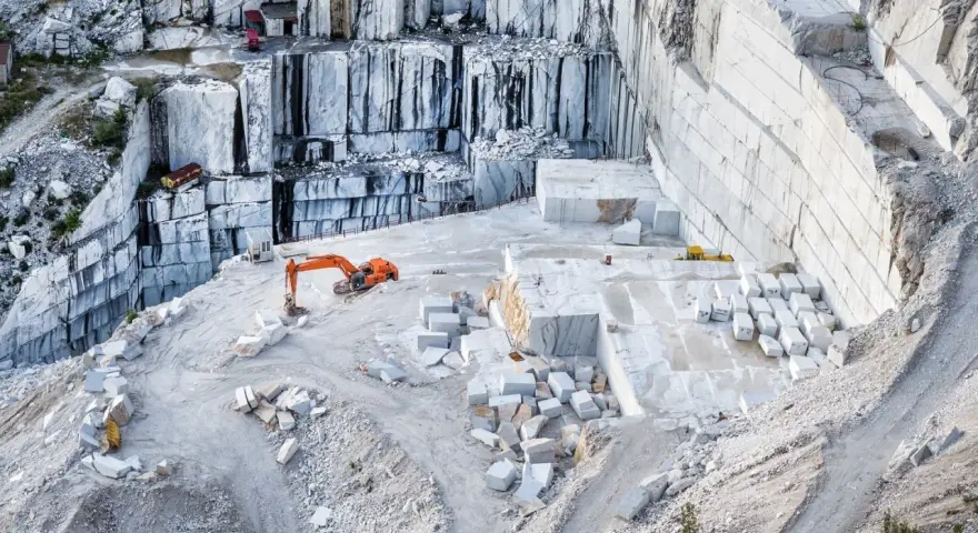 Cava di marmo a Carrara, con escavatore arancione al lavoro tra blocchi di pietra bianca. Un luogo affascinante da vedere a Carrara.