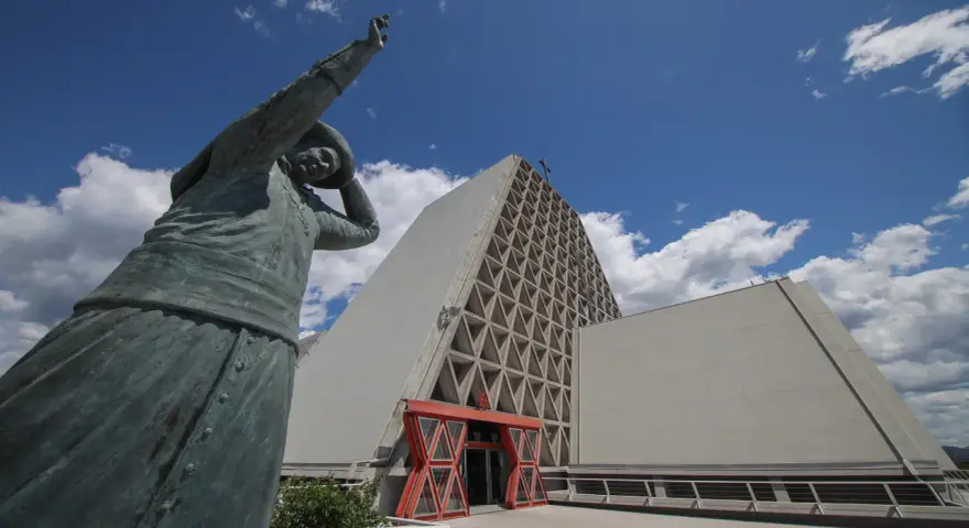 Statua bronzea davanti al Santuario Monte Grisa, con il suo ingresso rosso e la facciata a triangoli.