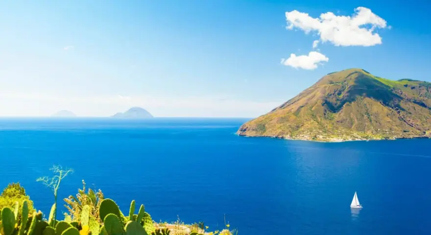 Panoramica delle Isole Eolie: cosa vedere? Un veliero solca il blu intenso del mare, con isole vulcaniche sullo sfondo e fichi d'India in primo piano.