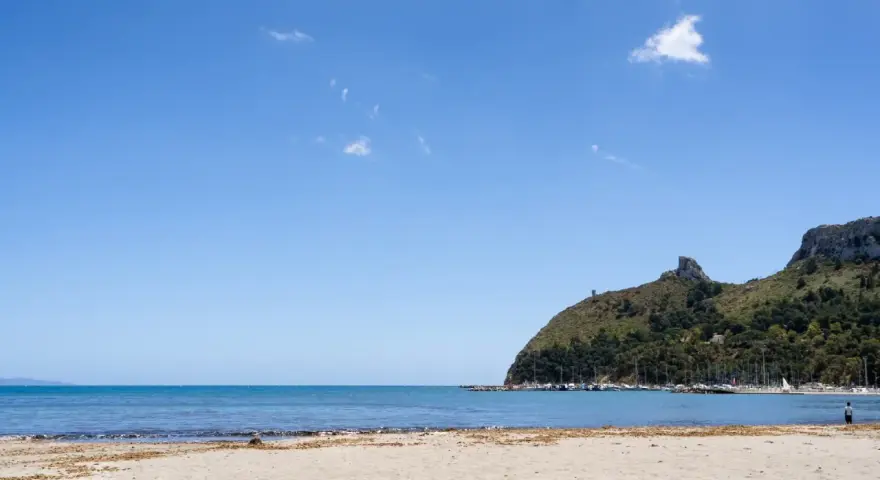 Spiaggia del Poetto con mare azzurro, montagne verdi e un porto turistico pieno di barche.