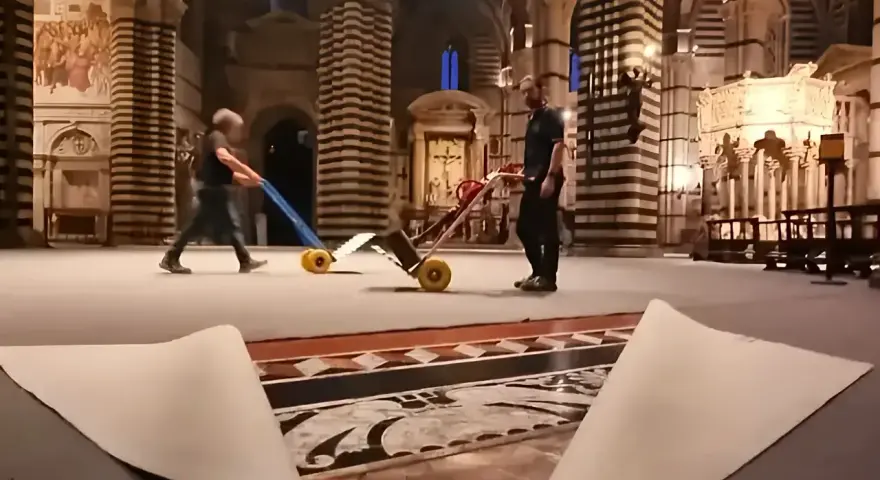 Due operai spostano carrelli sul magnifico pavimento del Duomo di Siena, mentre un telo protettivo è parzialmente sollevato, rivelando i dettagli artistici.