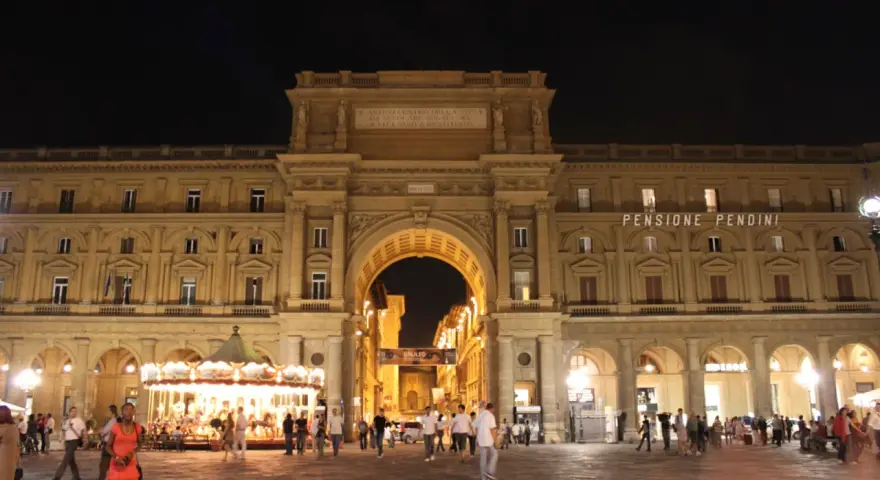 La piazza della Repubblica a Firenze di notte, con il suo arco monumentale illuminato, gente che passeggia e una giostra scintillante.