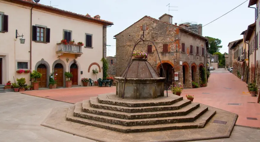 Piazza con pozzo antico a Civitella in Val di Chiana, circondata da case in pietra e intonaco, con fiori colorati.